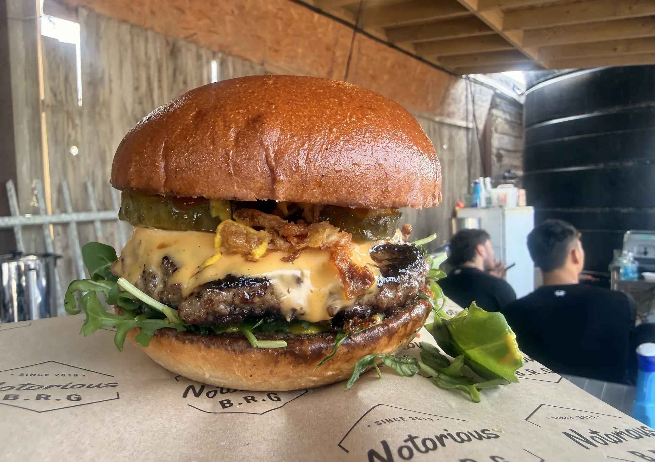 Double smash burger with cheese, pickles and crispy onions on Notorious B.R.G paper, with guests behind